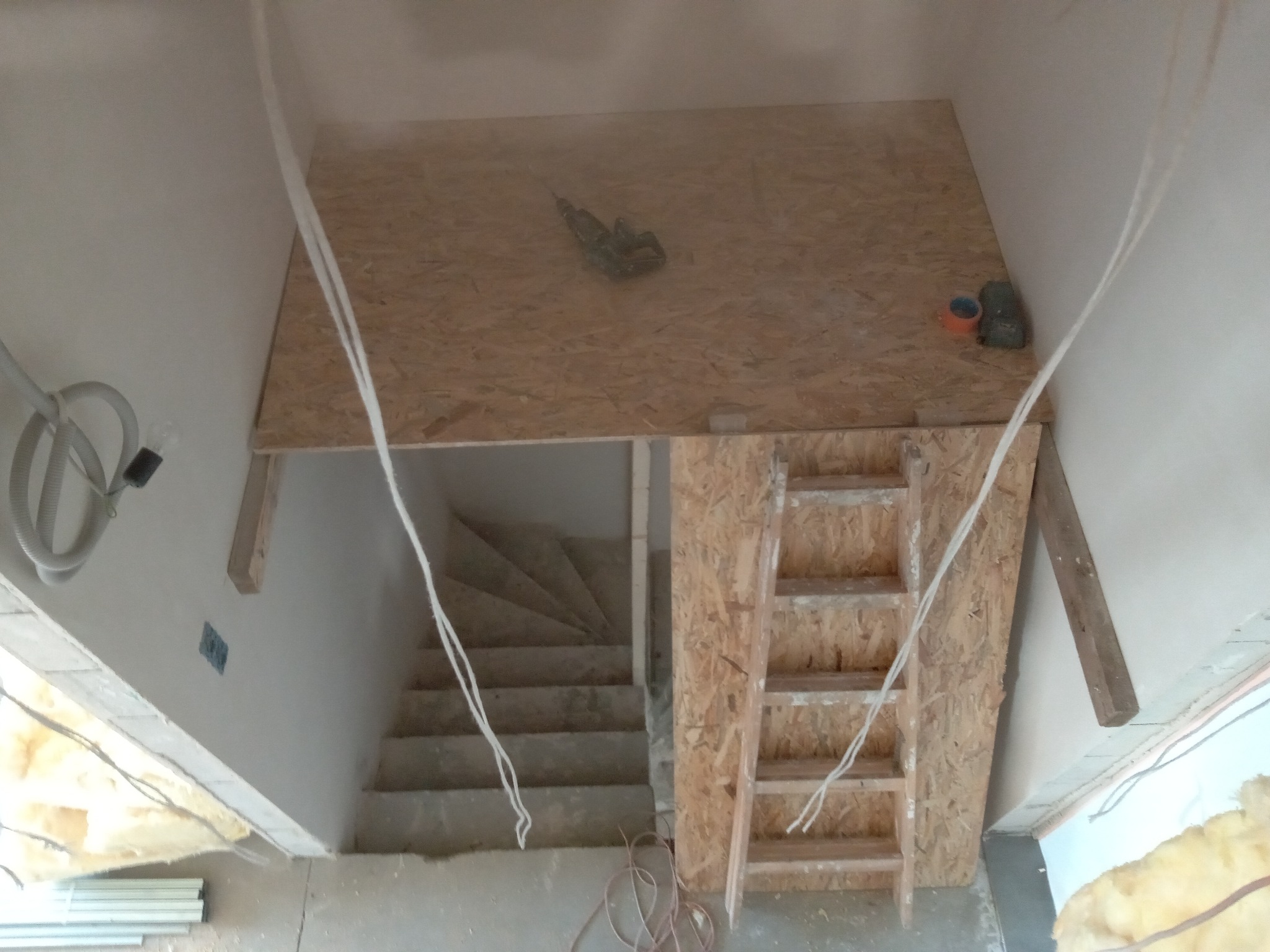Widok z góry na konstrukcję z płyty OSB nad schodami w trakcie prac, widoczne narzędzia, przewody elektryczne i drabina.
