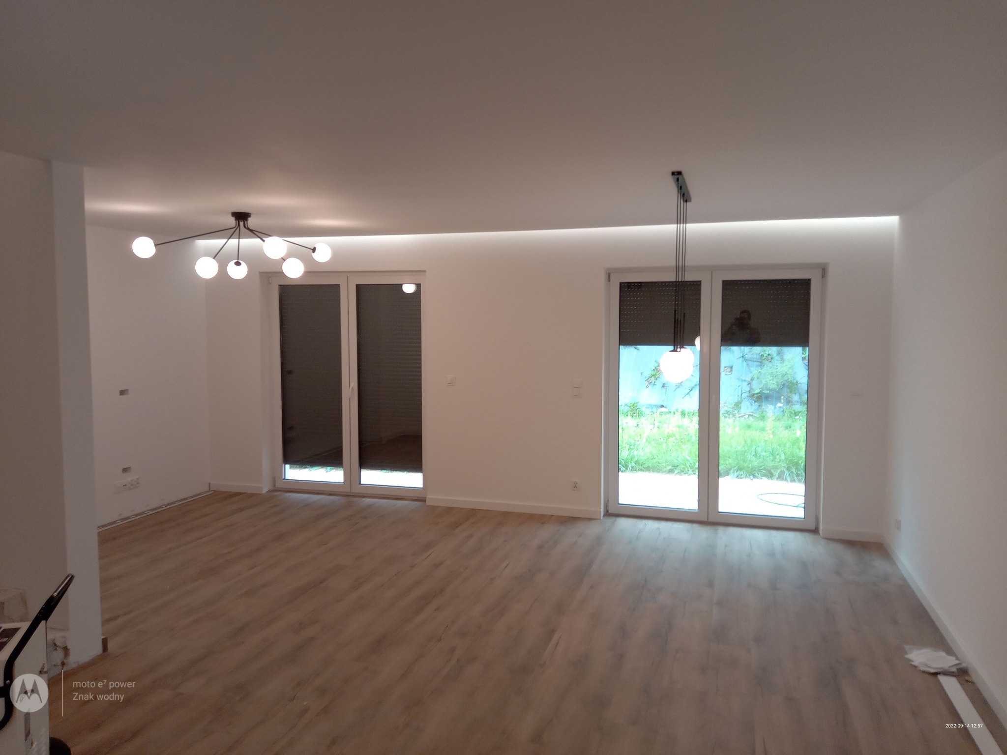 Jasne, przestronne wnętrze z panelami podłogowymi, oświetleniem LED w suficie podwieszanym i nowoczesnymi lampami wiszącymi, widok na zieleń przez duże okna balkonowe.