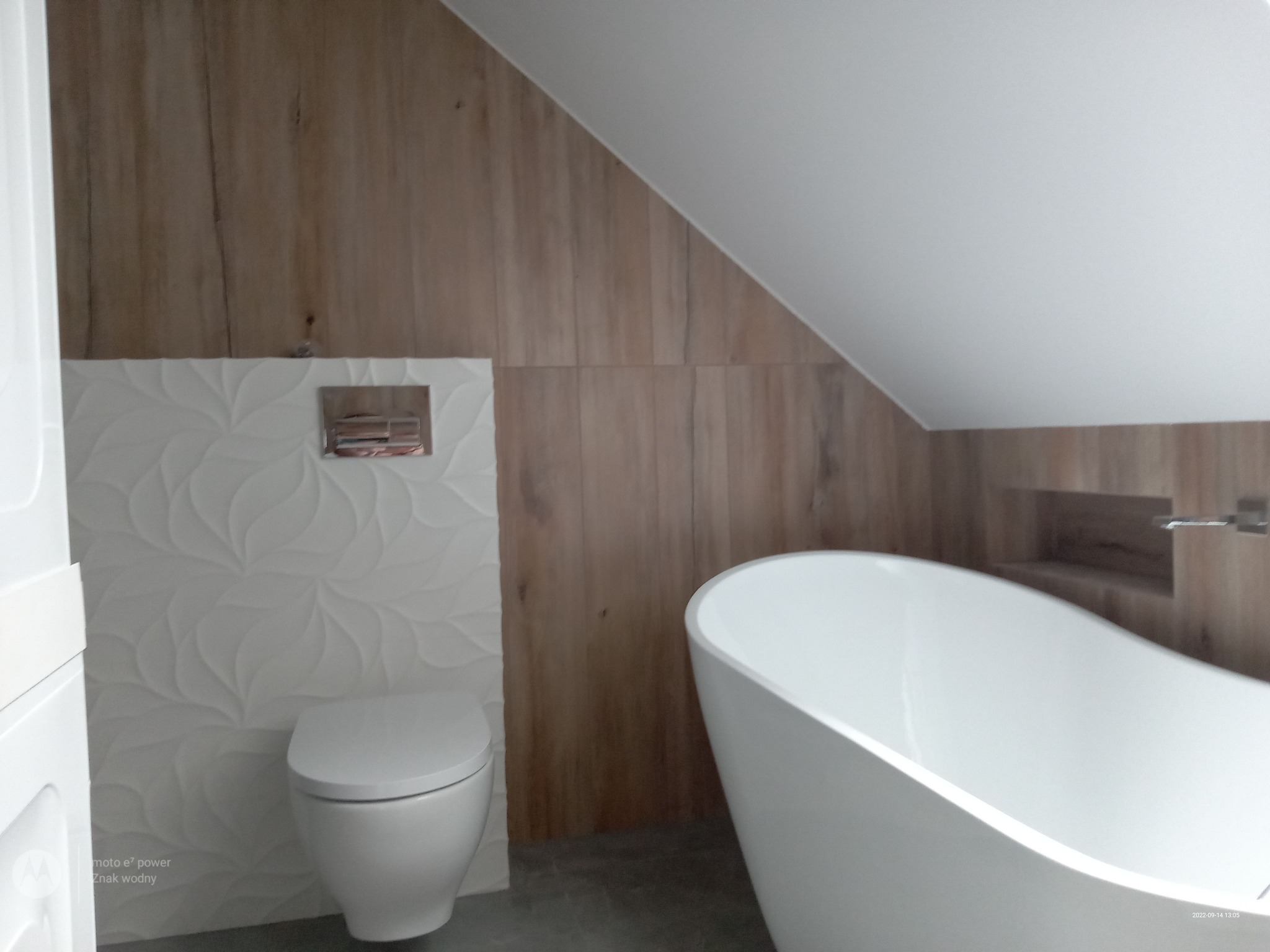 Nowoczesna łazienka na poddaszu z wolnostojącą białą wanną, toaletą i ścianą z białych, wzorzystych płytek oraz drewnopodobnych paneli ściennych.