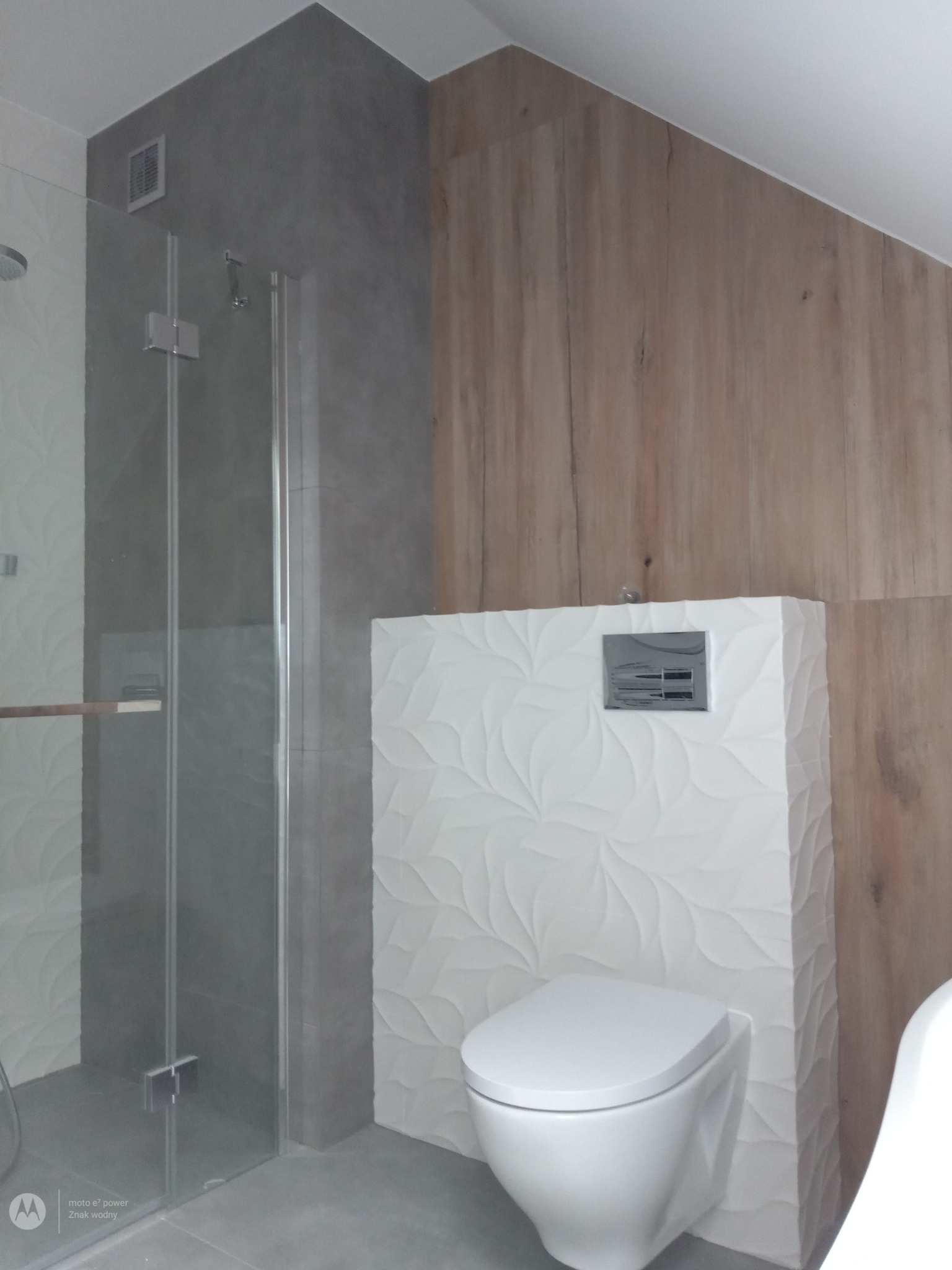 Nowoczesna łazienka z kabiną prysznicową ze szklanymi drzwiami, podwieszaną toaletą z deseniem liści na obudowie, szarą ścianą z wentylatorem i panelami drewnopodobnymi.