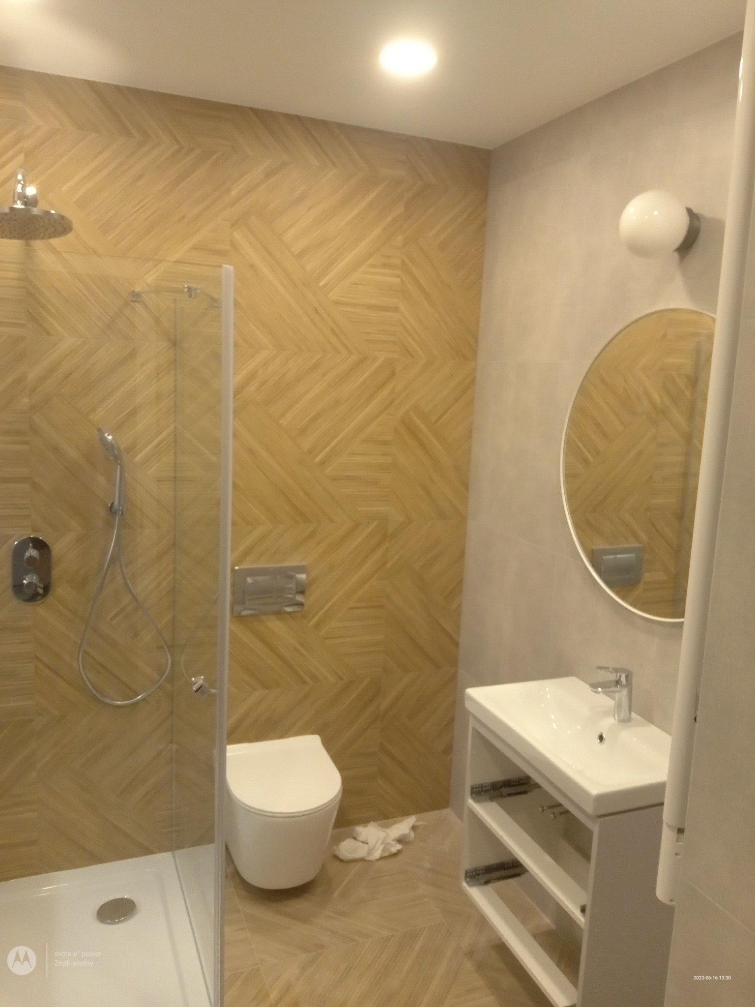 Nowoczesna łazienka z kabiną prysznicową, umywalką z szafką, okrągłym lustrem i toaletą w neutralnej kolorystyce z akcentem drewnopodobnej mozaiki na ścianach.