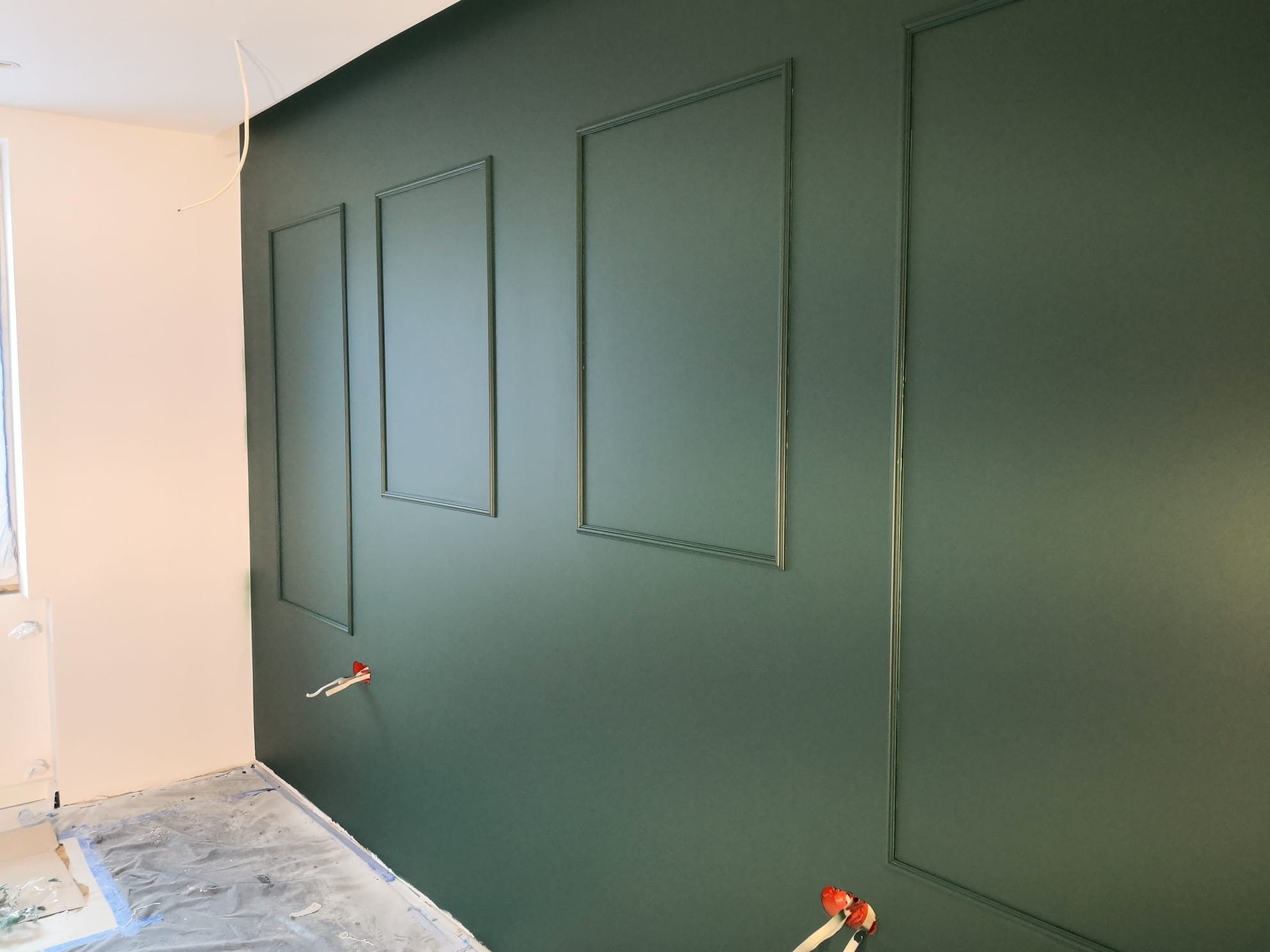 Ściana pomalowana na ciemnozielony kolor z ozdobnymi listwami w kształcie prostokątów, zabezpieczona folią podłoga, widoczne wystające kable elektryczne.