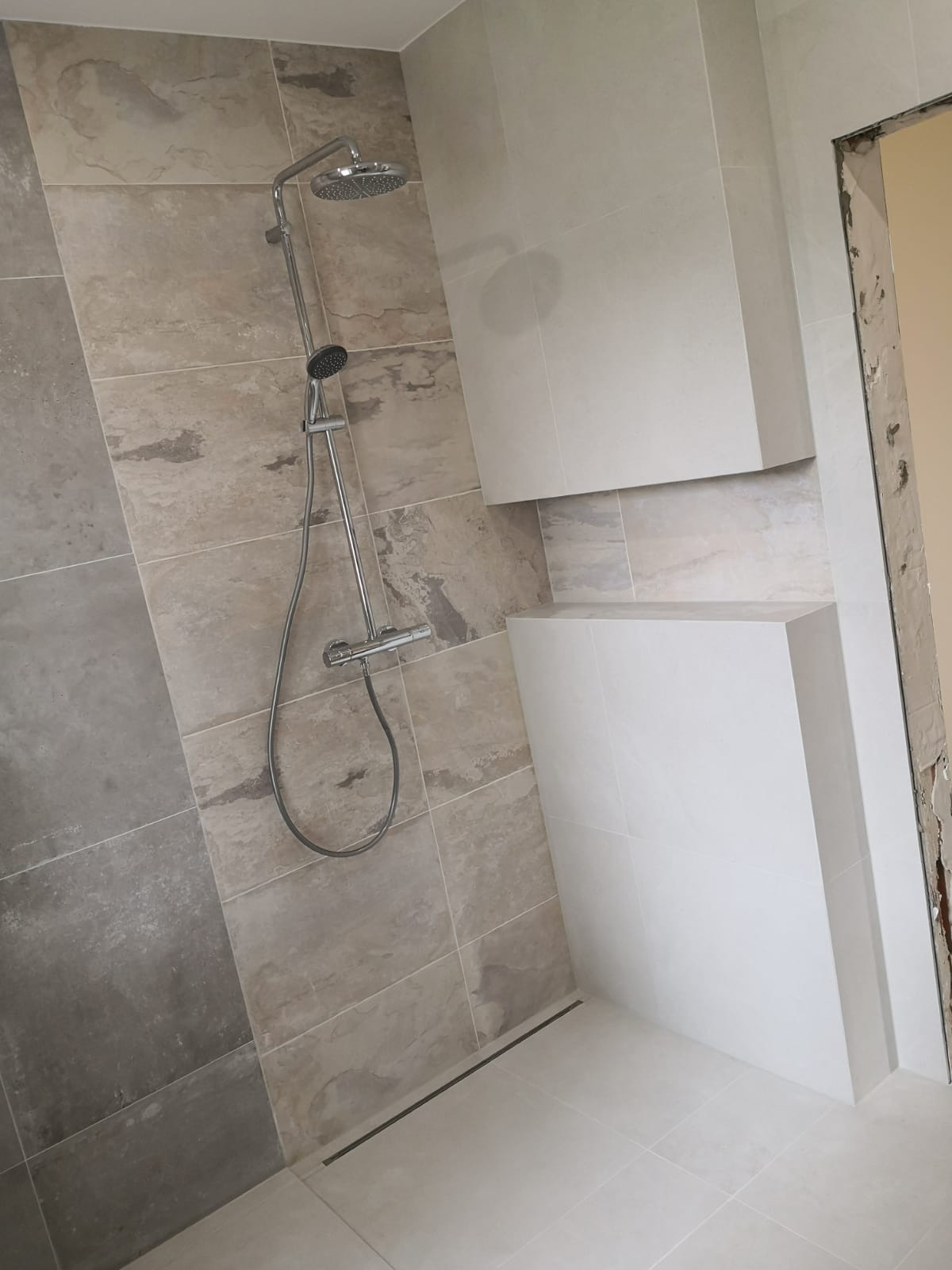 Nowoczesna kabina prysznicowa z szarymi płytkami imitującymi kamień, chromowaną armaturą i wbudowanymi półkami z białych płytek.