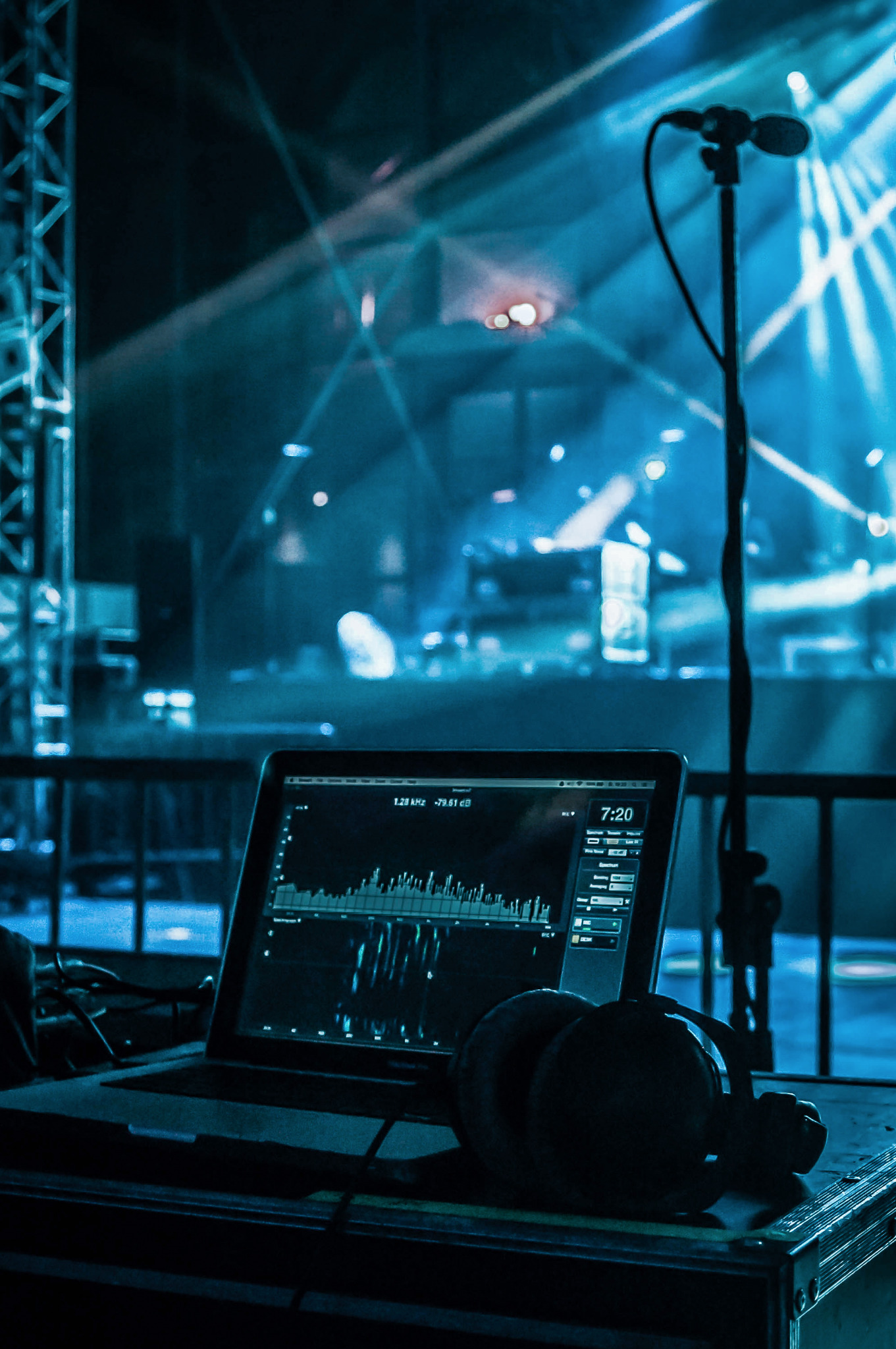 Laptop z widocznym oprogramowaniem do miksowania dźwięku, słuchawki leżące obok, mikrofon i oświetlenie sceniczne w tle, widok zza kulis.