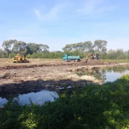Żółta ładowarka kołowa i zielona wywrotka z koparką na stercie ziemi przy powstającym stawie, widok zza bujnej roślinności.