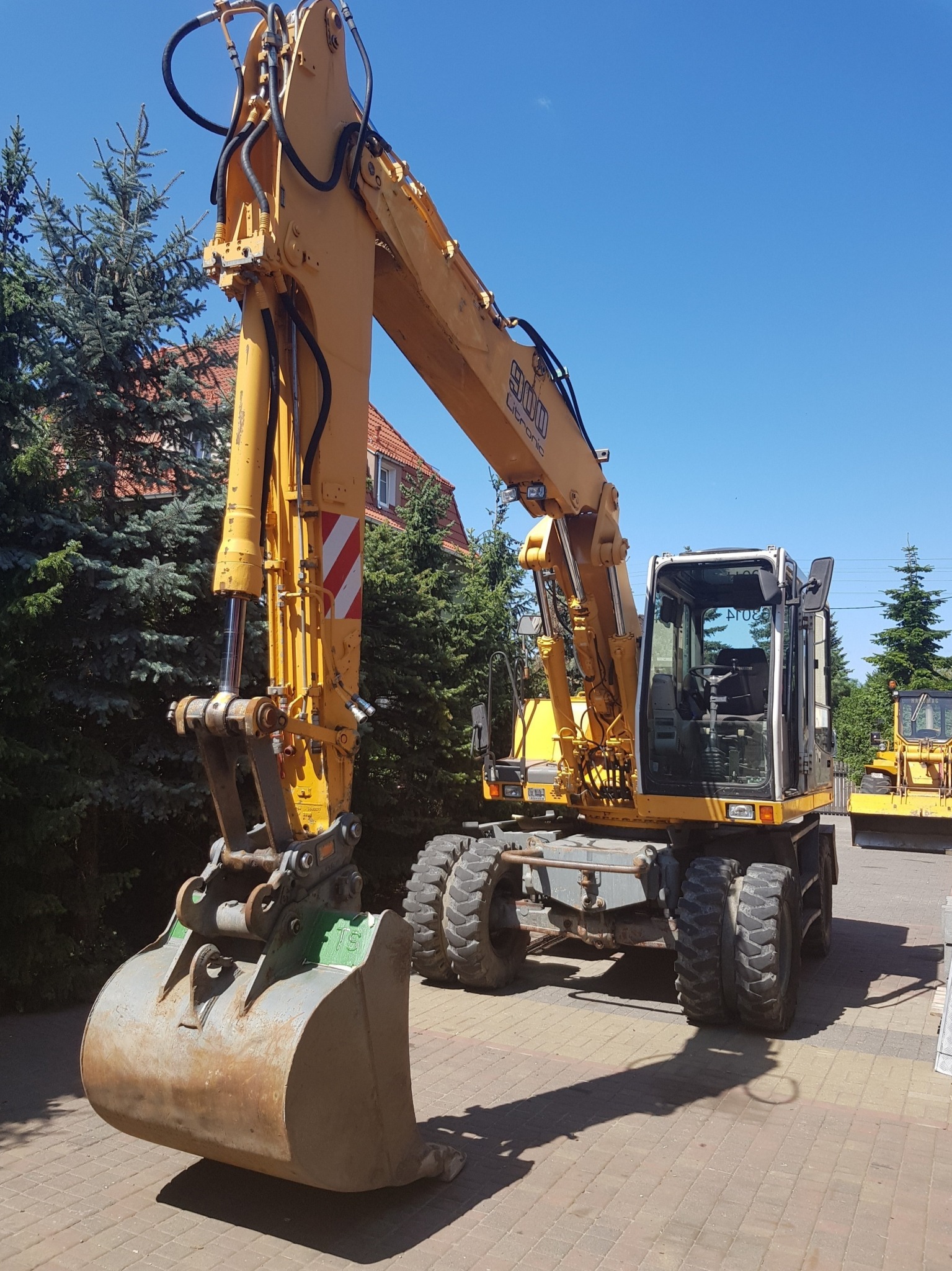 Żółta koparka kołowa Liebherr 900 Litronic z łyżką, stojąca na kostce brukowej, na tle zielonych drzew i błękitnego nieba. Widoczna inna maszyna budowlana w oddali.