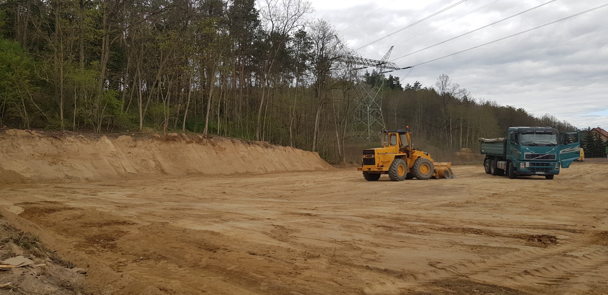 Żółta ładowarka kołowa i zielona wywrotka na placu budowy z usypaną pryzmą piasku, w tle las i linia wysokiego napięcia.