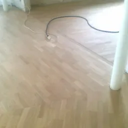 Parkiet ułożony w jodełkę, z widocznym przejściem do innego wzoru ułożenia desek, z przewodami elektrycznymi na podłodze i fragmentem kolumny w tle.