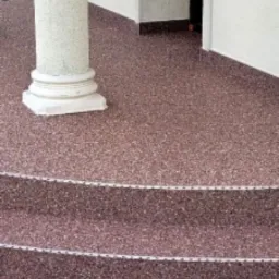 Wykończony taras z kolumną i schodami, pokryty czerwoną posadzką żywiczną z kamyczkami. Widoczna kostka brukowa na zewnątrz, białe ściany budynku.