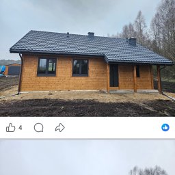 Imperial -Tim Adam Migo - Drewniany dom szkieletowy w Woli z ciemną dachówką, w stanie surowym zamkniętym. Widoczne okna i drzwi. Tło stanowią drzewa.