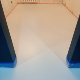 Imperial -Tim Adam Migo - Jasna, gładka posadzka żywiczna w pokoju, zabezpieczona taśmą malarską przy ścianach i progach. Fragmenty ciemnych, trójkątnych elementów po bokach kadru.