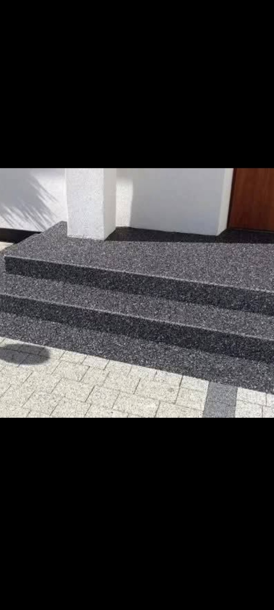 Zewnętrzne schody z posadzką żywiczną w kolorze grafitowym, prowadzące do drzwi wejściowych, z widoczną kostką brukową na podeście.