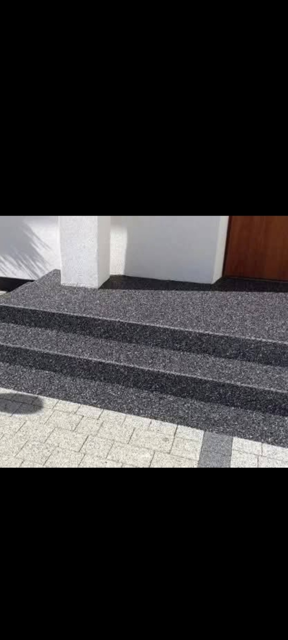 Zewnętrzne schody z szarą, drobnoziarnistą nawierzchnią, prowadzące do drzwi. Widoczny fragment chodnika z kostki brukowej.