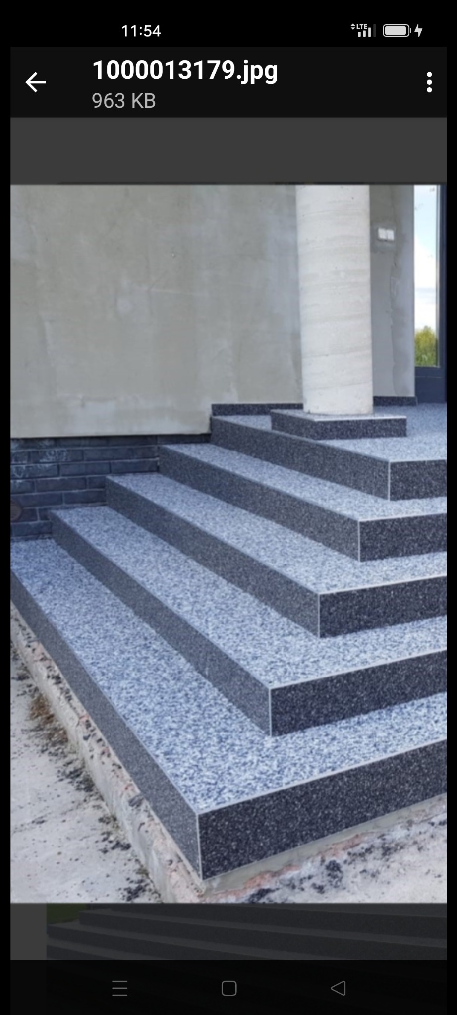 Zewnętrzne schody z szarego, drobnoziarnistego kamienia prowadzące do budynku. Widoczny fragment kolumny i ściana z surowym wykończeniem.