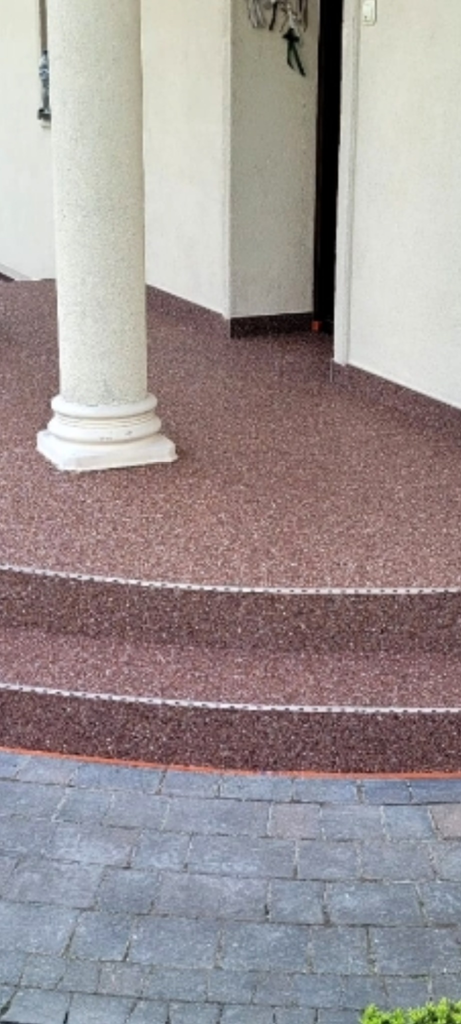 Wykończony taras z kolumną i schodami, pokryty czerwoną posadzką żywiczną z kamyczkami. Widoczna kostka brukowa na zewnątrz, białe ściany budynku.