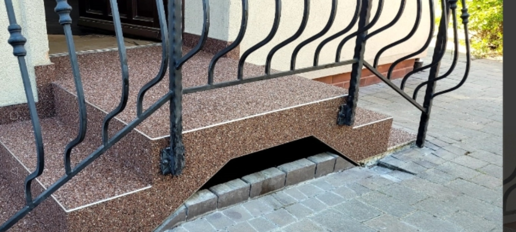 Zewnętrzne schody z żywicy w odcieniach brązu, wykończone białymi listwami, z czarną, metalową balustradą o dekoracyjnym wzorze, prowadzące do drzwi wejściowych.