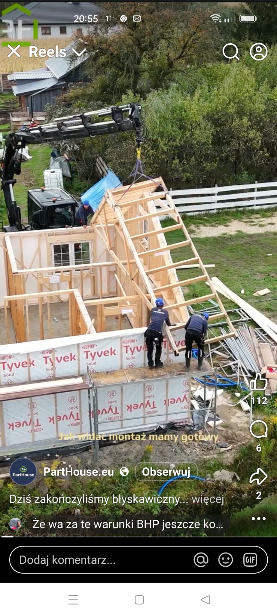 Montaż domu szkieletowego z użyciem dźwigu. Widoczni pracownicy w kaskach, elementy konstrukcyjne i membrany izolacyjne Tyvek. Ujęcie z zewnątrz, w tle zieleń.