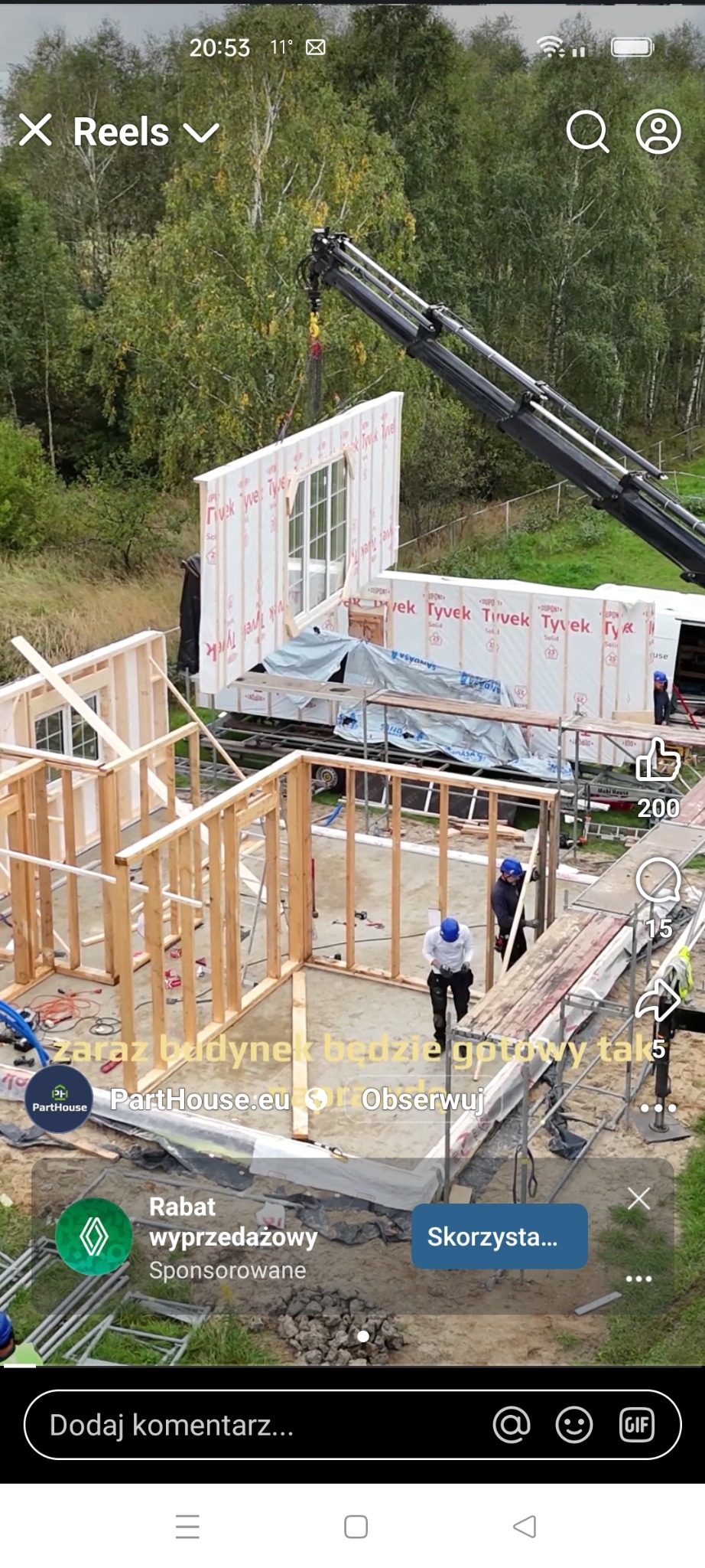 Montaż domu modułowego: dźwig unosi ścianę z oknem, widoczny szkielet konstrukcji i pracownicy w kaskach. Budowa w toku, elementy zabezpieczone folią Tyvek.