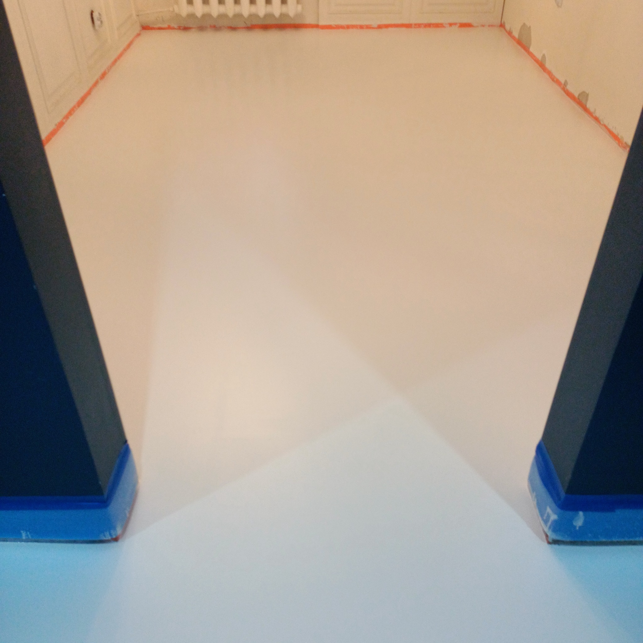 Jasna, gładka posadzka żywiczna w pokoju, zabezpieczona taśmą malarską przy ścianach i progach. Fragmenty ciemnych, trójkątnych elementów po bokach kadru.