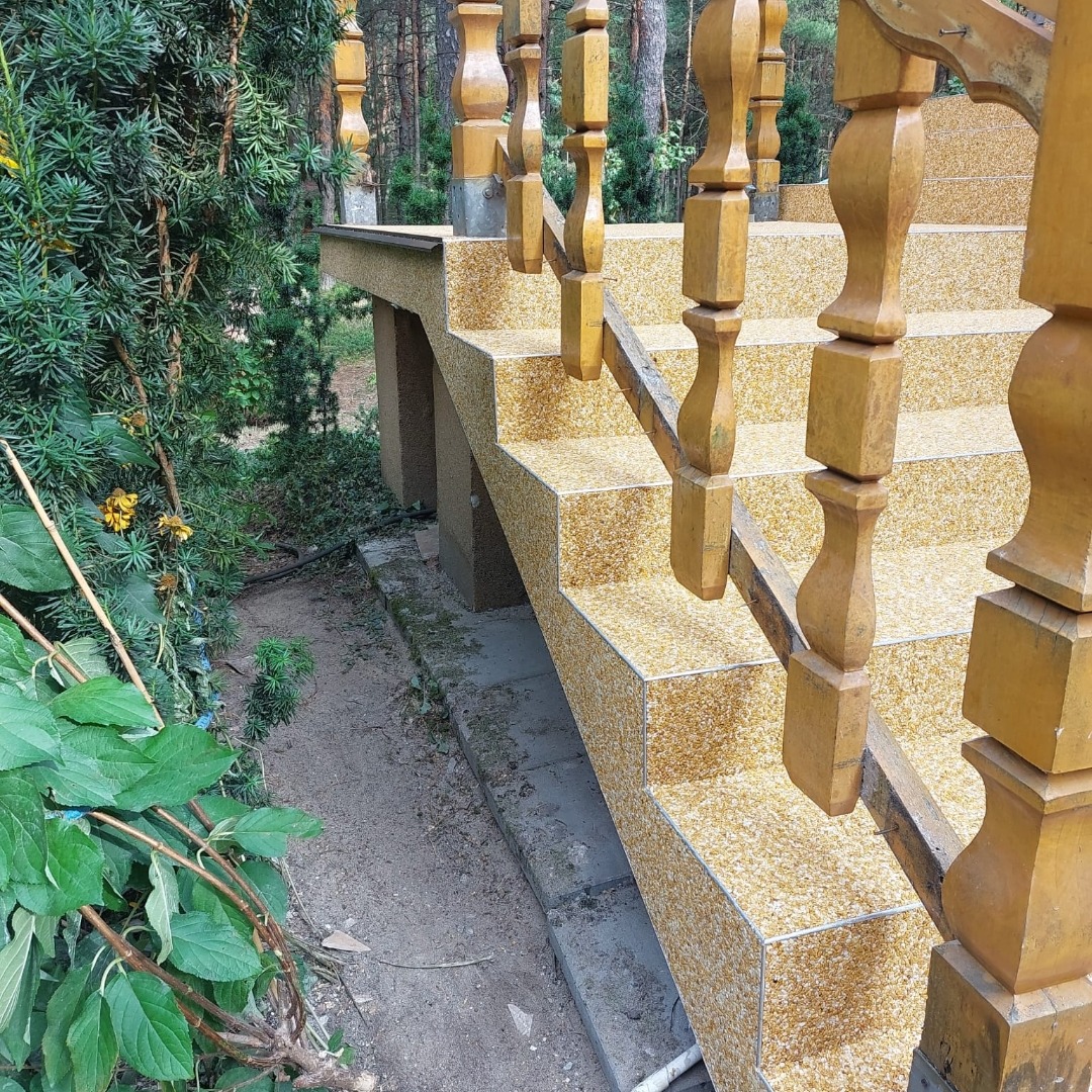 Zewnętrzne schody z balustradą, wykończone płytkami w odcieniach beżu i brązu, prowadzące do ogrodu. Drewniane elementy balustrady w ciepłym, żółtym odcieniu.