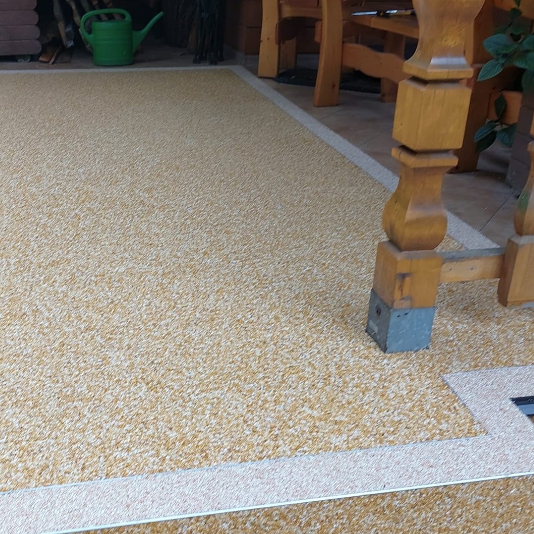 Beżowa posadzka żywiczna z delikatnym wzorem, obramowana jasnym paskiem, widoczna pod drewnianą konstrukcją z zieloną konewką w tle.