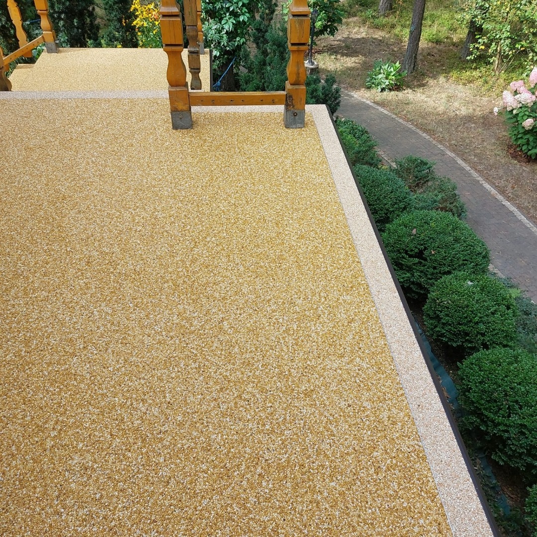 Żółta, drobnoziarnista posadzka żywiczna na tarasie z drewnianą balustradą, widok z góry na tle zieleni i ścieżki. Jasne, naturalne światło.