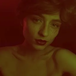 Portret kobiety z krótkimi włosami, czerwonymi ustami i brokatem na twarzy, oświetlonej czerwono-żółtym światłem na ciemnym tle.