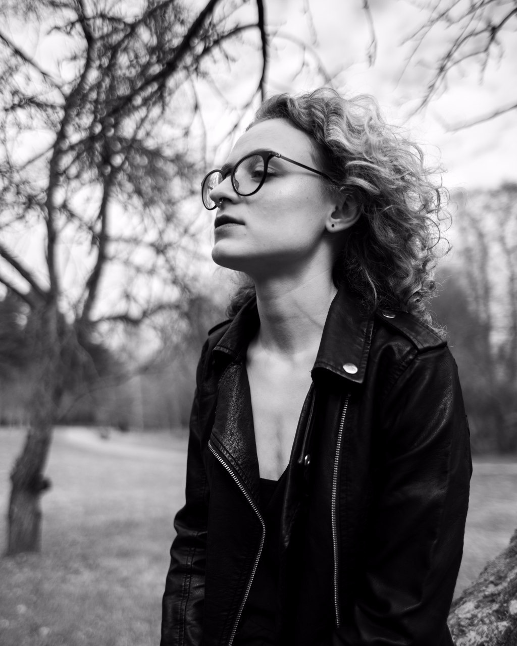 Czarno-białe zdjęcie kobiety w okularach, z kręconymi włosami i kolczykiem w nosie, ubrana w skórzaną kurtkę, spoglądająca w górę na tle drzew i rozmytego otoczenia.