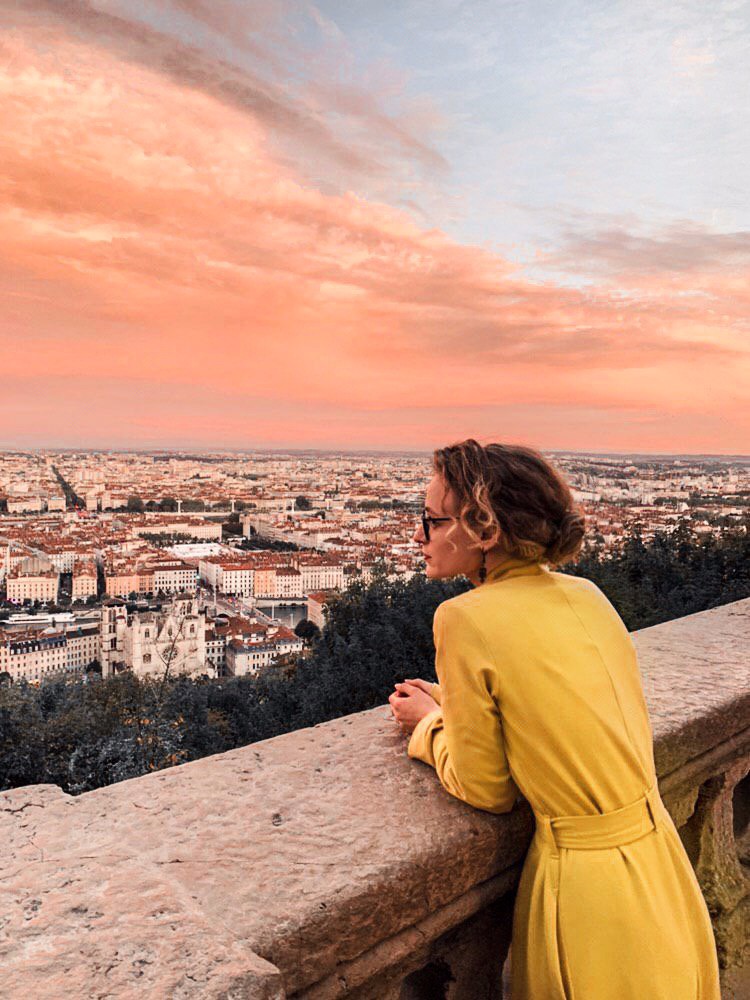 Kobieta w żółtym płaszczu zapiętym w pasie, opierająca się o kamienną balustradę z widokiem na miasto w oddali i różowe chmury o zachodzie słońca.