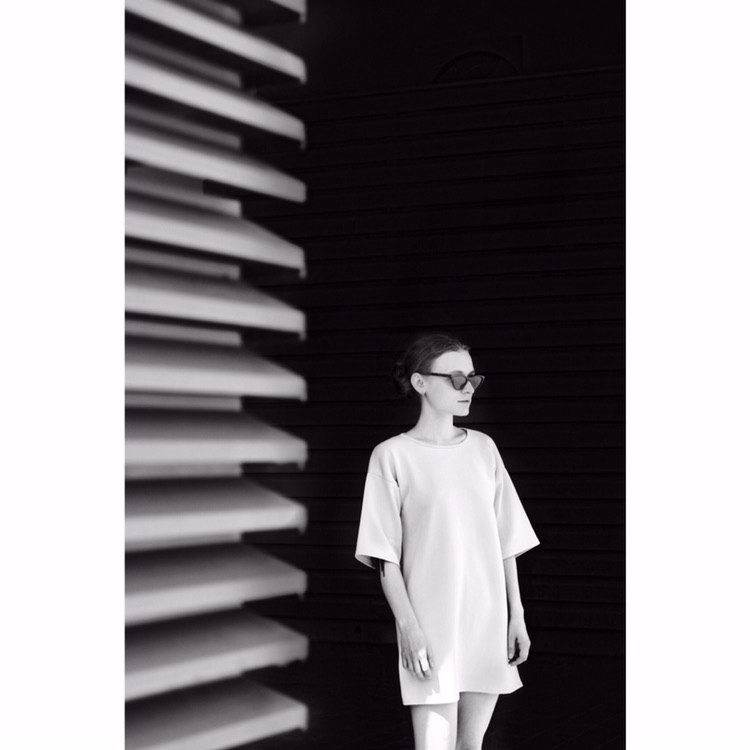 Czarno-białe zdjęcie kobiety w okularach przeciwsłonecznych, stojącej przed ścianą z poziomymi panelami, oświetlonej z lewej strony przez światło padające przez żaluzje.