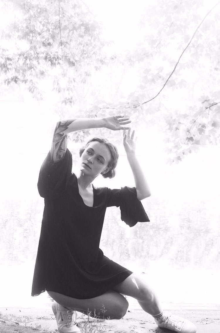 Czarno-białe zdjęcie kobiety w sukience, kucającej z podniesionymi rękami, tatuaż na ramieniu, tło z drzewami.