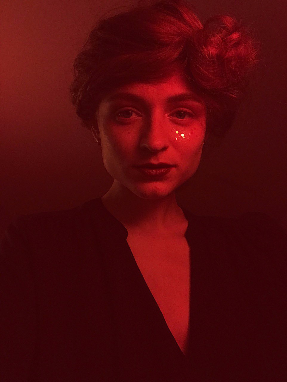 Portret kobiety w czerwonym oświetleniu, z błyszczącymi elementami makijażu pod okiem, ubrana w ciemną bluzkę z dekoltem w serek.