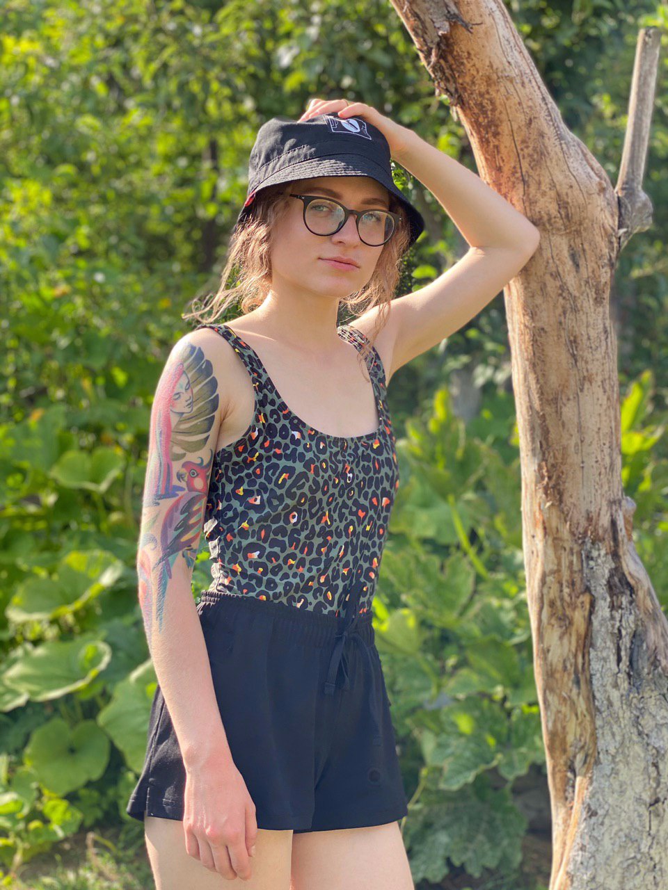 Kobieta w okularach i kapeluszu typu bucket hat, opierająca się o uschnięte drzewo, z tatuażem na ramieniu, w letnim stroju na tle zieleni.