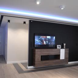 Firma BUD-JACK - Nowoczesne wnętrze: czarna ściana z telewizorem, drewniana szafka RTV z białym akcentem, podwieszany sufit z niebieskim oświetleniem LED, panele podłogowe w odcieniach szarości i beżu, widoczne...