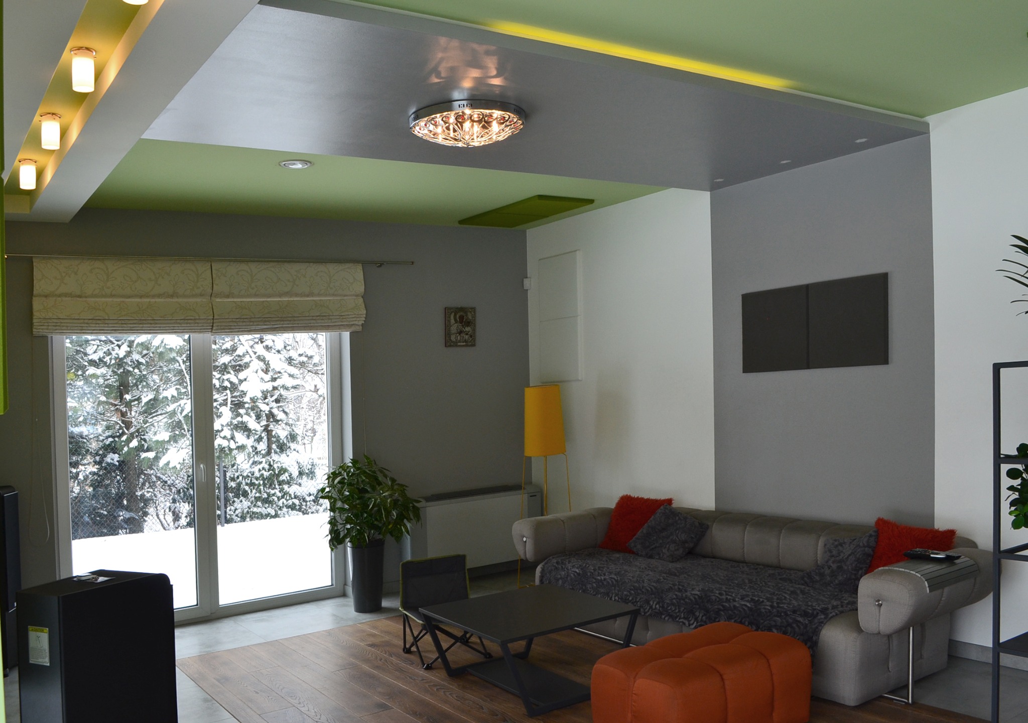 Nowoczesny salon z drewnianą podłogą, szarą sofą z pomarańczowymi poduszkami, widokiem na zaśnieżony ogród przez duże okno i wielobarwnym sufitem.