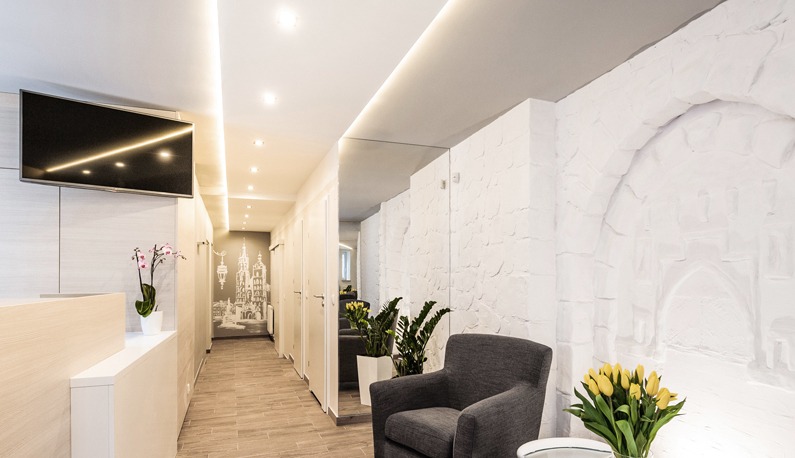 Nowoczesne wnętrze przedpokoju z dekoracyjną ścianą o fakturze kamienia, oświetleniem LED w suficie, telewizorem na ścianie i elementami dekoracyjnymi jak kwiaty i grafika na ścianie.