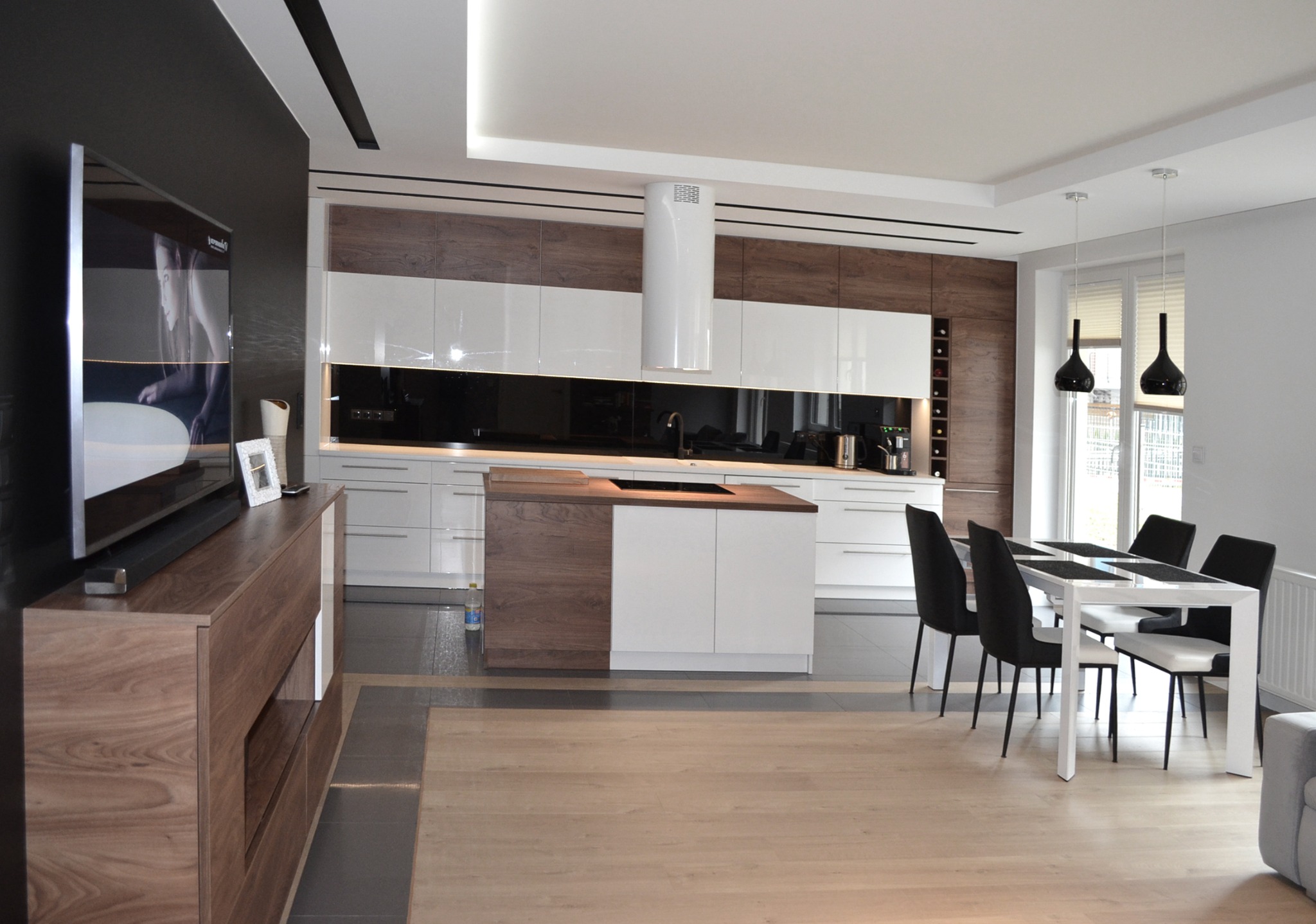 Nowoczesna kuchnia z wyspą, biało-drewniane szafki, czarny panel nad blatem, stół z czarnymi krzesłami, podłoga z jasnych paneli.