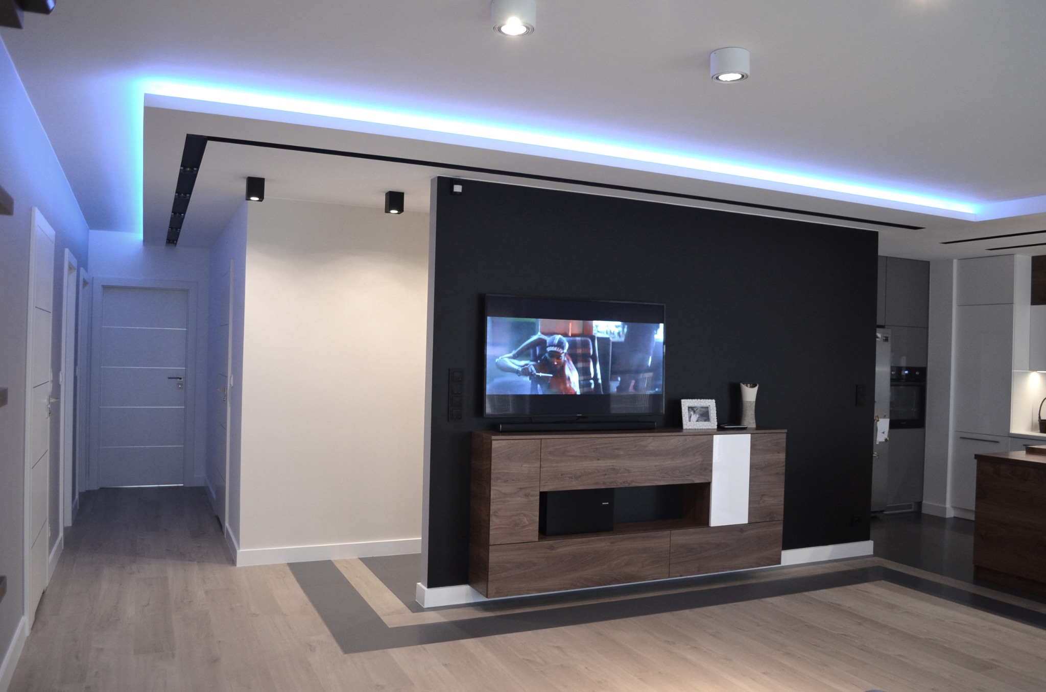 Nowoczesne wnętrze: czarna ściana z telewizorem, drewniana szafka RTV z białym akcentem, podwieszany sufit z niebieskim oświetleniem LED, panele podłogowe w odcieniach szarości i beżu, widoczne...