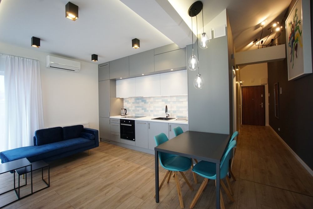 Nowoczesne wnętrze mieszkania z otwartą kuchnią, drewnianą podłogą, szarymi szafkami kuchennymi z białymi frontami i turkusowymi krzesłami przy czarnym stole.