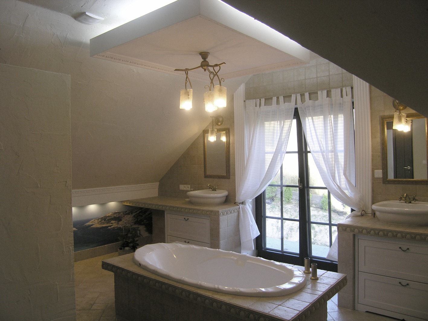 Wykończona łazienka na poddaszu z wanną na podwyższeniu, umywalkami nablatowymi, oknem balkonowym z białymi zasłonami i oświetleniem punktowym.