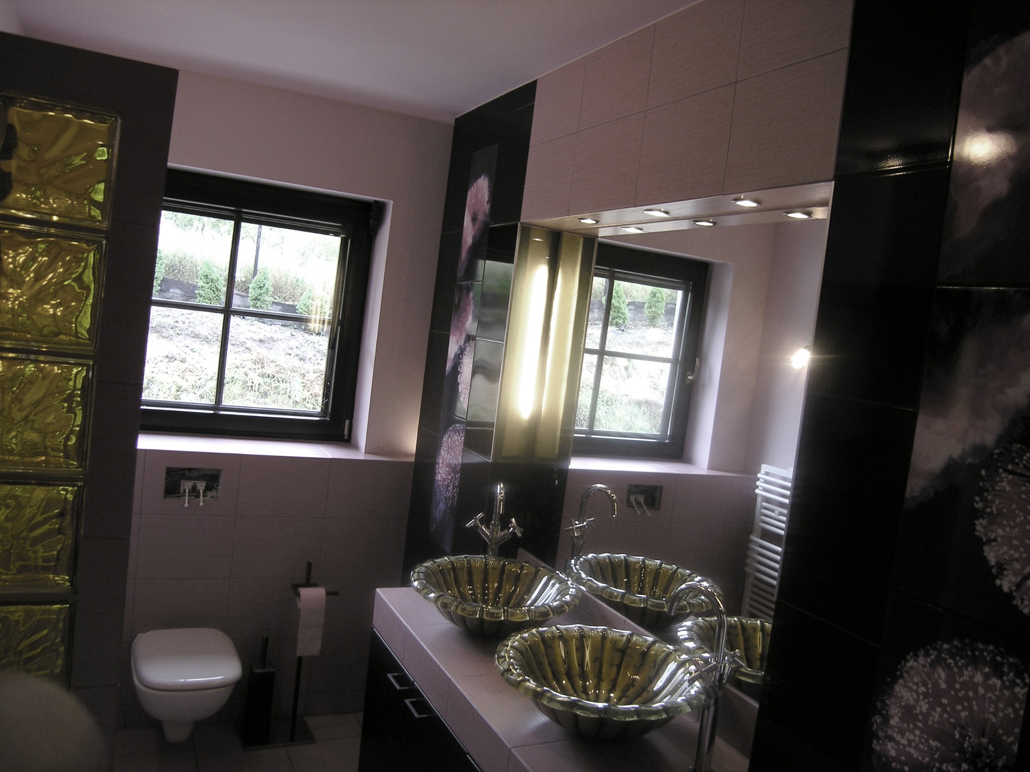 Nowoczesna łazienka z dwoma dekoracyjnymi umywalkami w kształcie muszli, czarnymi płytkami z motywem kwiatowym, oknem z widokiem na zieleń i toaletą w tle.