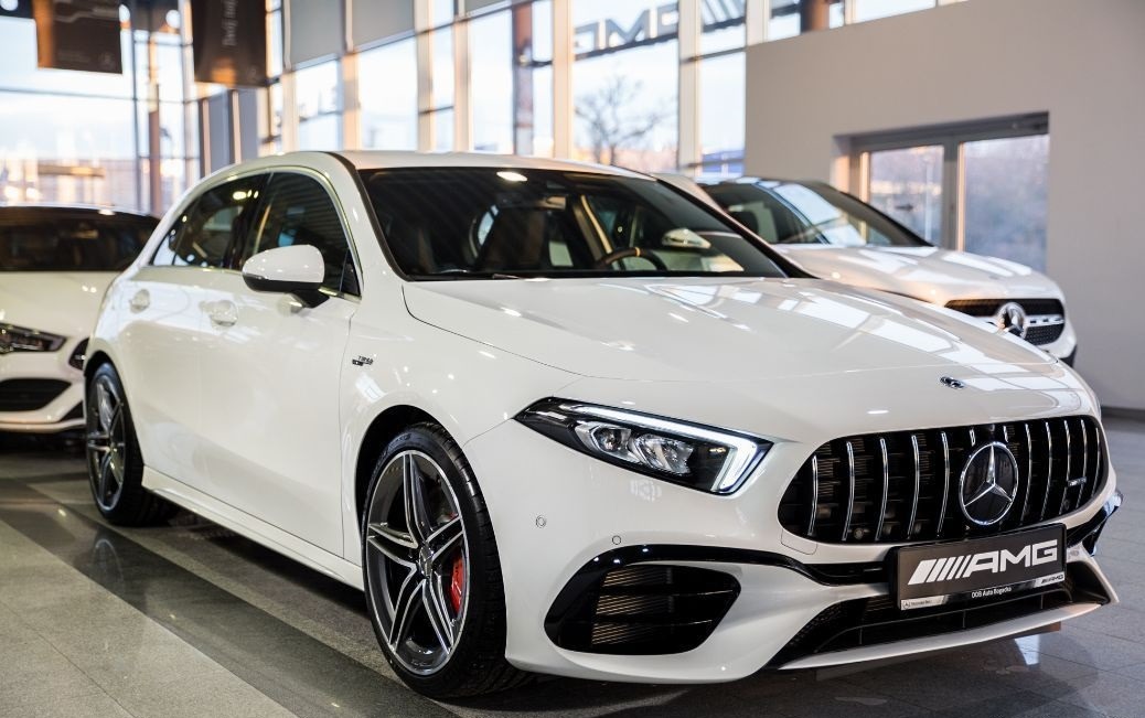 Biały samochód marki Mercedes-Benz AMG A-klasa, widok z przodu, z charakterystycznym grillem AMG i felgami z czerwonymi zaciskami hamulcowymi, na tle salonu samochodowego.