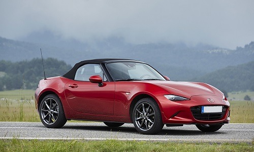 Czerwony kabriolet Mazda MX-5 z czarnym dachem, zaparkowany na drodze z zielonym poboczem i zalesionymi wzgórzami w tle.