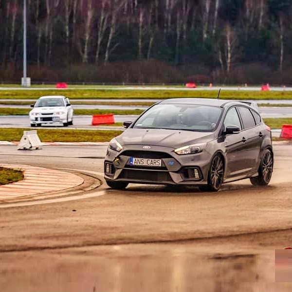 Szary Ford Focus RS na torze wyścigowym, widoczny z przodu, z tablicą rejestracyjną 'ANS CARS', w tle inny samochód i drzewa.