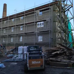 Budynek w trakcie prac elewacyjnych, widoczne rusztowania, sterta materiałów budowlanych, miniładowarka Bobcat i konstrukcja stalowa w tle.