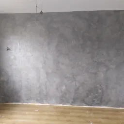 Ściana w pokoju po nałożeniu dekoracyjnego tynku w odcieniach szarości, widoczne ślady po gniazdku elektrycznym, podłoga drewniana.