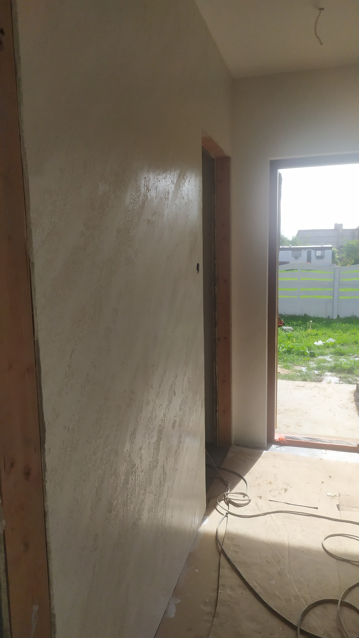 Ściana z nałożoną, dekoracyjną, jasną strukturą w trakcie prac wykończeniowych, widoczne kable na podłodze i surowa ościeżnica drzwiowa, w tle widok na ogród.