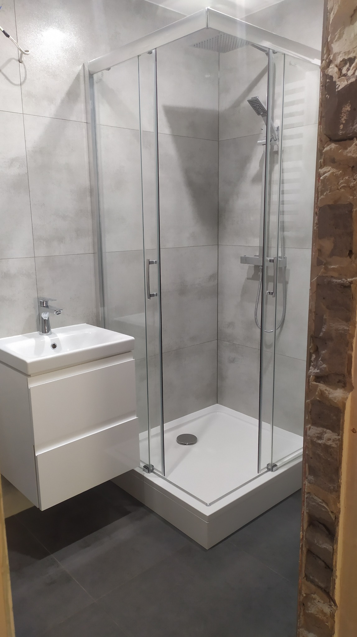 Nowoczesna łazienka z kabiną prysznicową narożną, białą umywalką z szafką podwieszaną, szarymi płytkami ściennymi imitującymi beton i fragmentem ściany z surowej cegły.