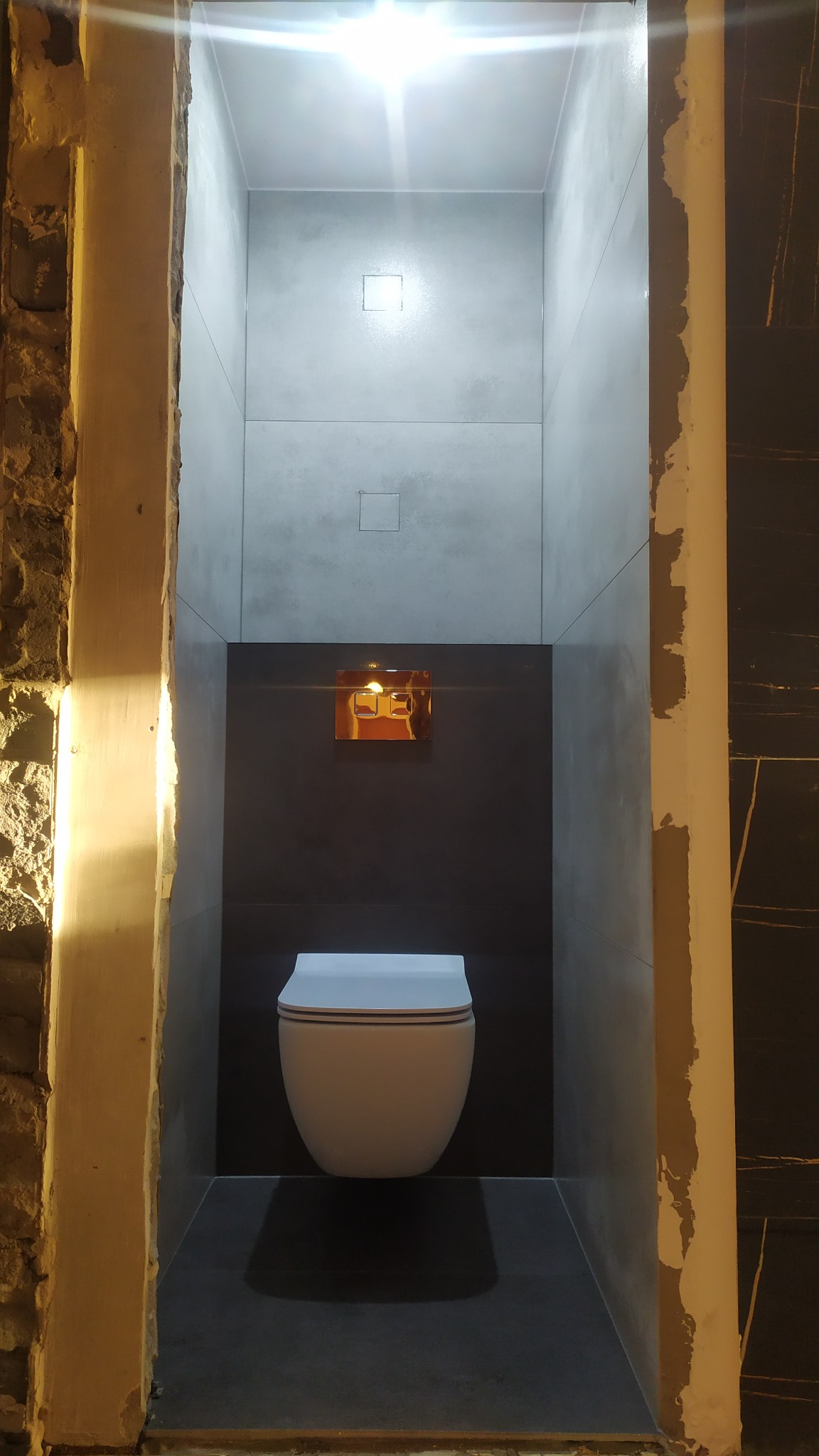 Nowoczesna toaleta wisząca w wąskiej wnęce wyłożonej szarymi płytkami, z widocznym fragmentem surowej ściany po lewej stronie i czarną płytką z żyłami po prawej, oświetlona górnym światłem.