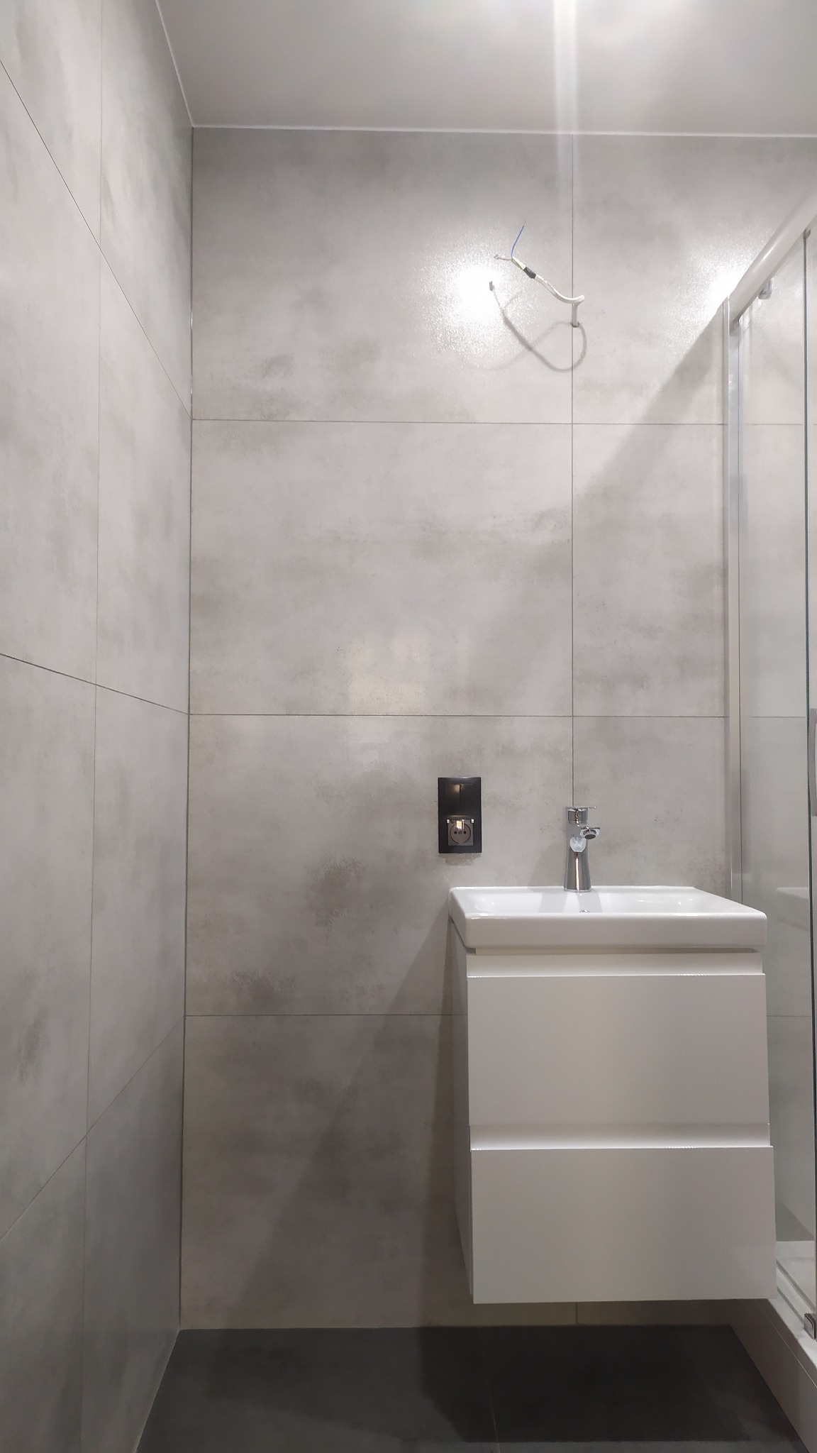 Nowoczesna łazienka z szarymi, betonopodobnymi płytkami, białą szafką pod umywalkę i czarną podłogą z płytek. Widoczne niezakończone podłączenie elektryczne. Minimalistyczny styl.